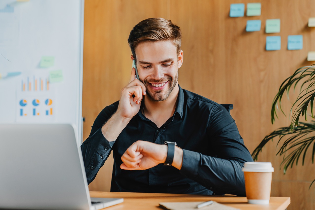 jovem empresário olhando para relógio para ver a Best Time to Call, o melhor horário para ligar para o cliente, com celular na mão