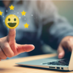 A imagem mostra um close de uma pessoa interagindo com um computador. O foco está em um dedo tocando a tela ou gesticulando em direção a um emoji sorridente, cercado por cinco estrelas douradas. O emoji e as estrelas sugerem satisfação ou uma avaliação positiva, enquanto o laptop no fundo reforça um contexto digital ou online, provavelmente relacionado a feedback ou experiência do cliente. A composição passa uma sensação de positividade e interação tecnológica.