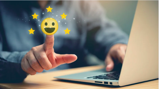 A imagem mostra um close de uma pessoa interagindo com um computador. O foco está em um dedo tocando a tela ou gesticulando em direção a um emoji sorridente, cercado por cinco estrelas douradas. O emoji e as estrelas sugerem satisfação ou uma avaliação positiva, enquanto o laptop no fundo reforça um contexto digital ou online, provavelmente relacionado a feedback ou experiência do cliente. A composição passa uma sensação de positividade e interação tecnológica.