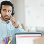 A imagem mostra um profissional de atendimento ao cliente usando um headset, indicando que ele está envolvido em uma conversa ou suporte remoto. Ele parece estar explicando algo, com uma expressão séria e engajada. Em primeiro plano, há outro profissional de costas, também com um headset, sugerindo um ambiente de call center ou suporte técnico. Ao fundo, é possível ver um quadro branco desfocado, reforçando um cenário de trabalho colaborativo.