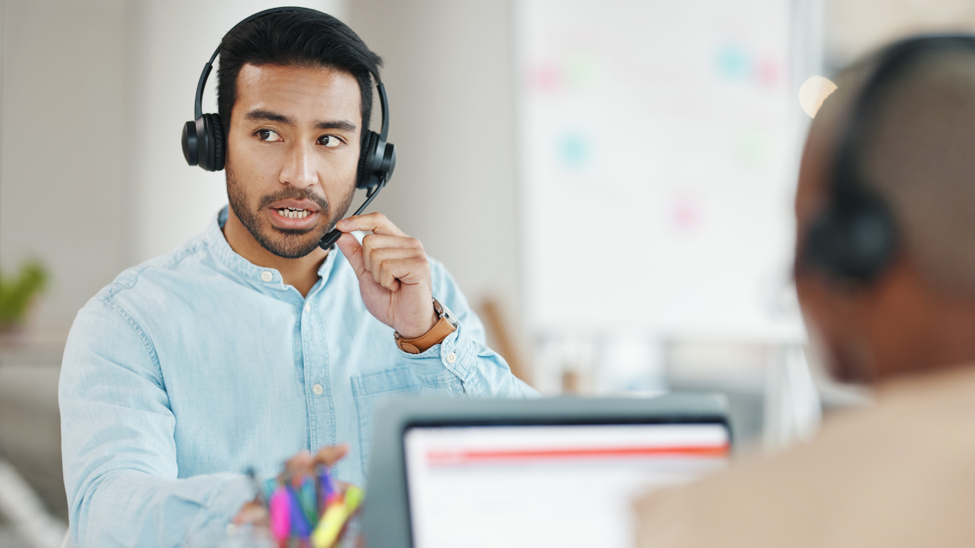 A imagem mostra um profissional de atendimento ao cliente usando um headset, indicando que ele está envolvido em uma conversa ou suporte remoto. Ele parece estar explicando algo, com uma expressão séria e engajada. Em primeiro plano, há outro profissional de costas, também com um headset, sugerindo um ambiente de call center ou suporte técnico. Ao fundo, é possível ver um quadro branco desfocado, reforçando um cenário de trabalho colaborativo.